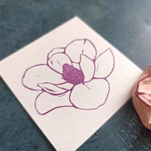 Może przedstawiać: Zbliżenie na gumową pieczątkę z detalicznym wzorem magnolii. Pieczątka jest r&oacute;żowa, a kwiat jest obwiedziony fioletowym kolorem.