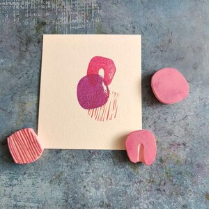 May include: A cream-colored card with a pink and purple abstract design, accompanied by three pink rubber stamps. The stamps have various shapes, including a circle, a curved shape, and a textured block. The background is a textured blue surface.