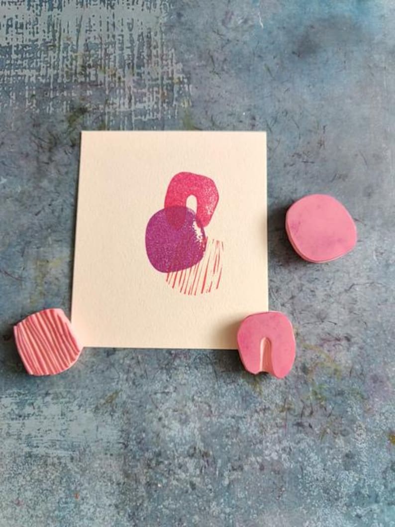May include: A cream-colored card with a pink and purple abstract design, accompanied by three pink rubber stamps. The stamps have various shapes, including a circle, a curved shape, and a textured block. The background is a textured blue surface.