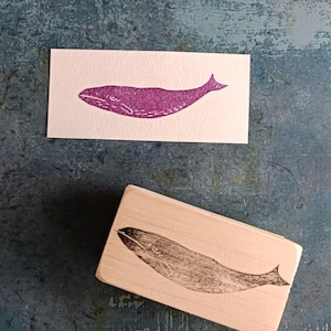 May include: A wooden block stamp with a whale design. The whale is carved into the wood and is a dark gray color. The stamp is on a light blue surface.