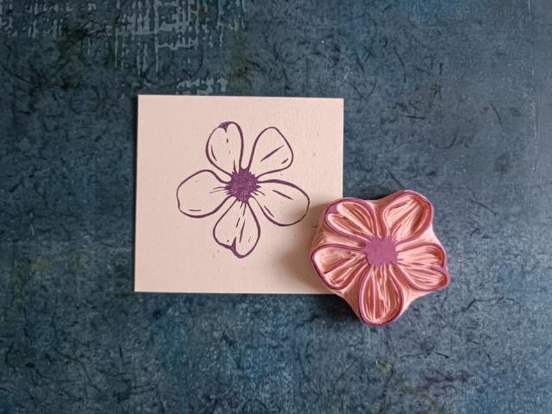 Pode incluir: Um carimbo de borracha rosa com um design de flor detalhado. O carimbo &eacute; usado para criar uma impress&atilde;o de flor roxa em um cart&atilde;o rosa claro.