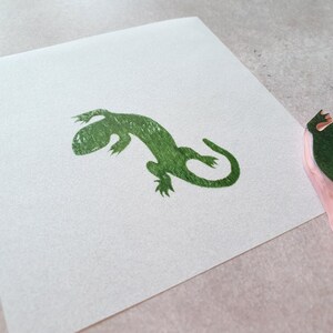 May include: A green lizard stamp next to a matching print on white paper. The stamp has a pink base. The lizard print is a detailed silhouette, perfect for crafting or art projects.