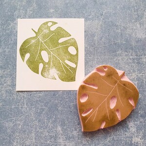 May include: A green monstera leaf stamp next to a print of the same leaf. The stamp is brown with pink details. The leaf print is on a white square. The background is a textured blue.