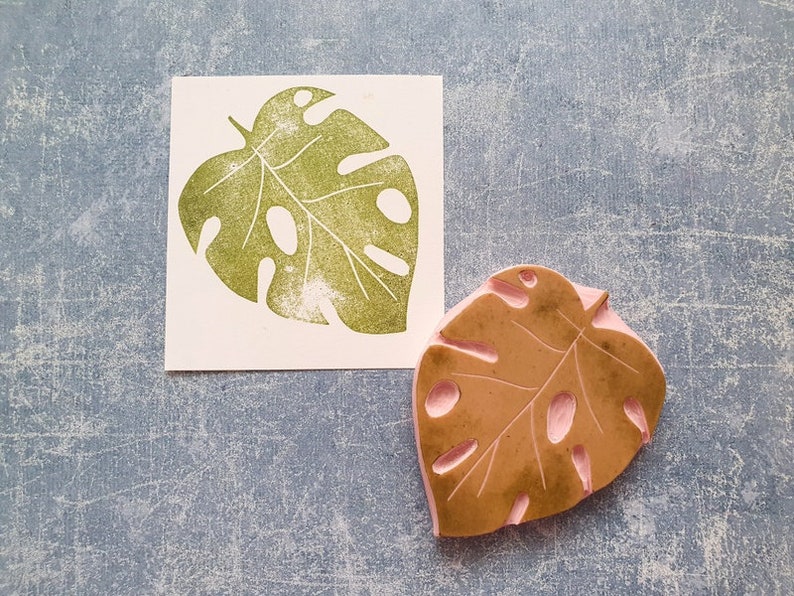 May include: A green monstera leaf stamp next to a print of the same leaf. The stamp is brown with pink details. The leaf print is on a white square. The background is a textured blue.