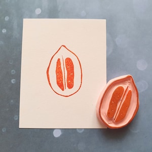 May include: A pink rubber stamp with a red outline of a halved fruit with two seeds inside. The stamp is on a white card with a red impression of the same fruit.