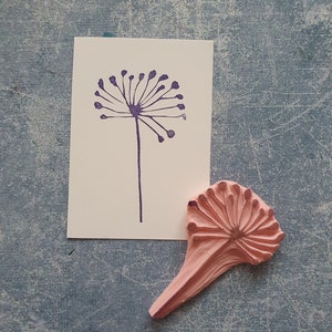 May include: A pink rubber stamp with a detailed design of a dandelion flower. The stamp is on a light blue textured surface.