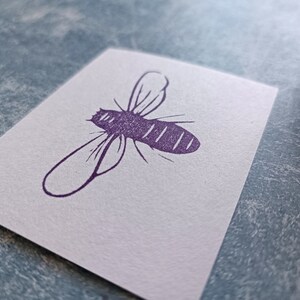 May include: A white card with a purple bee illustration. The bee is depicted in a simple, stylized design with wings and a segmented body. The card is on a textured, light blue surface.