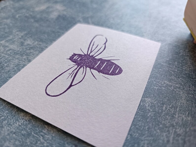 May include: A white card with a purple bee illustration. The bee is depicted in a simple, stylized design with wings and a segmented body. The card is on a textured, light blue surface.
