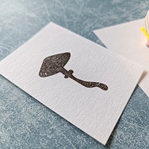 K&ouml;nnte beinhalten: Ein schwarzer Gummistempel mit einem Pilzmotiv. Der Stempel befindet sich auf einer wei&szlig;en Karte.