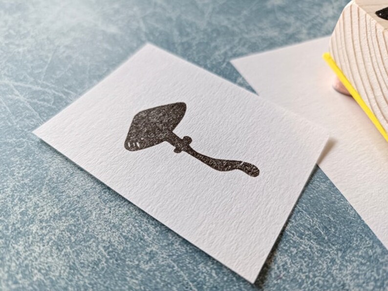 K&ouml;nnte beinhalten: Ein schwarzer Gummistempel mit einem Pilzmotiv. Der Stempel befindet sich auf einer wei&szlig;en Karte.