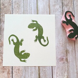 May include: A green lizard stamp next to a print of two lizards. The stamp is pink with a black lizard design. The lizards are printed in green ink on a white square of paper. The background is a light brown wooden surface.