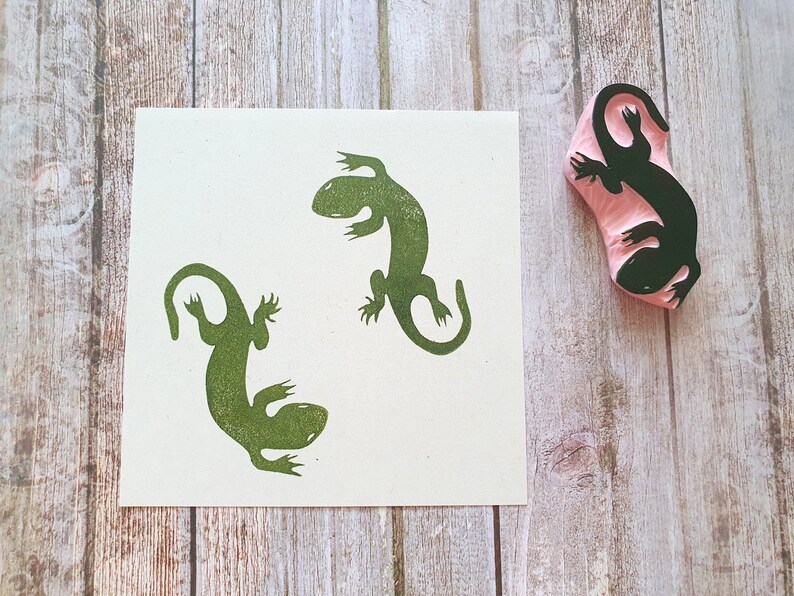 May include: A green lizard stamp next to a print of two lizards. The stamp is pink with a black lizard design. The lizards are printed in green ink on a white square of paper. The background is a light brown wooden surface.