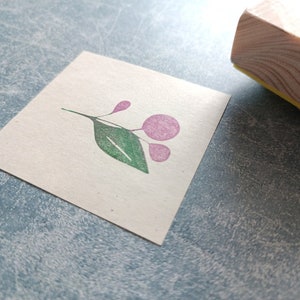 以下が含まれることがあります： 白いカードにピンクと緑の花柄のゴム印。印は木製の台に載っている。