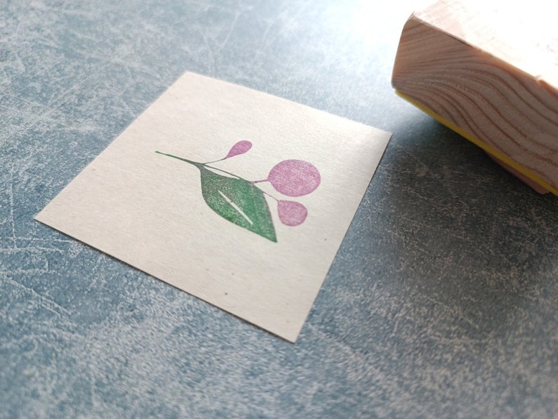 以下が含まれることがあります： 白いカードにピンクと緑の花柄のゴム印。印は木製の台に載っている。