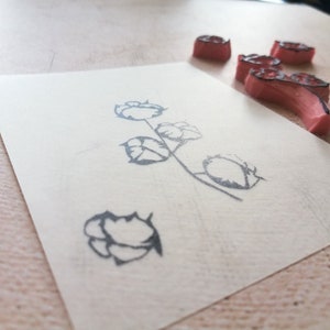 May include: A floral design stamp with a cotton plant image on a piece of paper. The stamp is made of pink rubber with a black outline. The paper is off-white, and the background is a light wood surface.