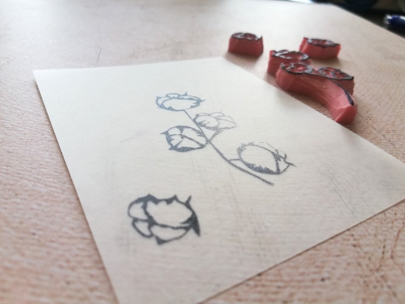 May include: A floral design stamp with a cotton plant image on a piece of paper. The stamp is made of pink rubber with a black outline. The paper is off-white, and the background is a light wood surface.