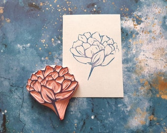 May include: A pink rubber stamp with a detailed floral design of a peony flower. The stamp is on a blue background with gold flecks. A white card with a blue ink impression of the same floral design is next to the stamp.