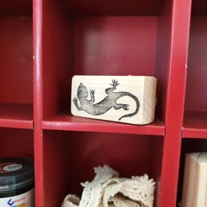 May include: A wooden stamp featuring a detailed black lizard design. The stamp is rectangular and sits inside a red cubby shelf. The shelf also contains a jar of black paint and other craft supplies.