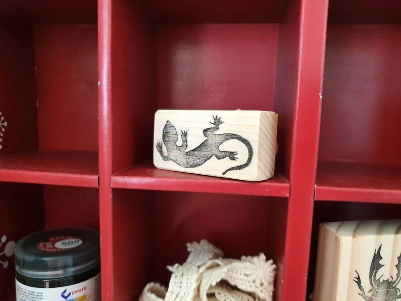 May include: A wooden stamp featuring a detailed black lizard design. The stamp is rectangular and sits inside a red cubby shelf. The shelf also contains a jar of black paint and other craft supplies.
