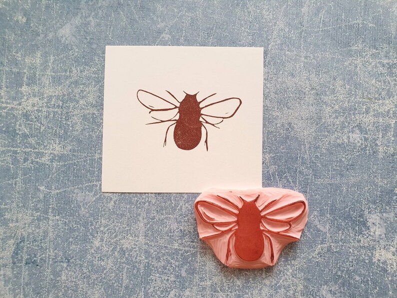 May include: A brown insect stamp and its impression on white paper. The stamp is made of pink rubber and features a detailed insect design. The paper is placed on a textured blue surface.