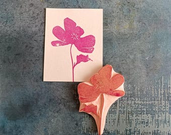 May include: A pink flower stamp and its impression on a white card. The stamp is carved from wood and shows a detailed floral design. The card displays a vibrant pink floral print, showcasing the stamp's intricate details. The background is a textured blue surface.