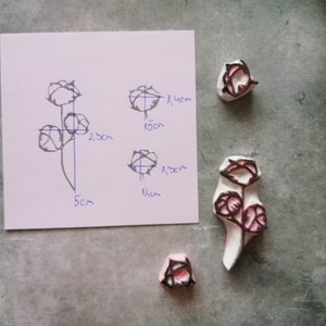 May include: A collection of handmade rose stamps and a sketch. The stamps are white with pink and black rose details. The sketch on a white paper shows a rose design with measurements in centimeters.