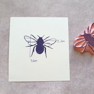 May include: A bee stamp and its impression. The stamp is pink with a purple bee design. The bee impression is dark blue on a white square, with dimensions of 3.6 cm by 5.2 cm, or approximately 1.4 inches by 2 inches.