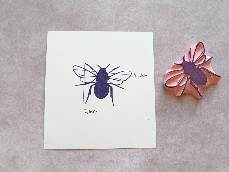 May include: A bee stamp and its impression. The stamp is pink with a purple bee design. The bee impression is dark blue on a white square, with dimensions of 3.6 cm by 5.2 cm, or approximately 1.4 inches by 2 inches.