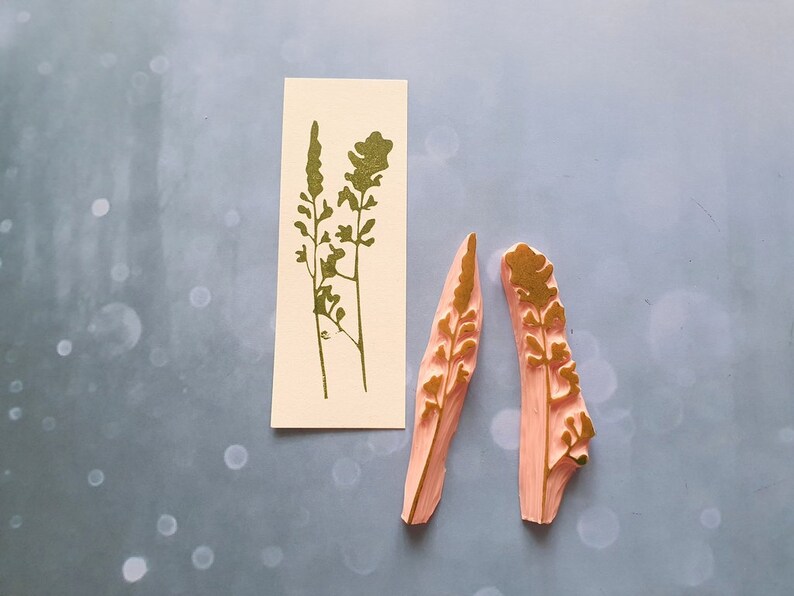 May include: Two pink rubber stamps with gold leaf designs of stylised floral sprigs. The stamps are on a light blue background.