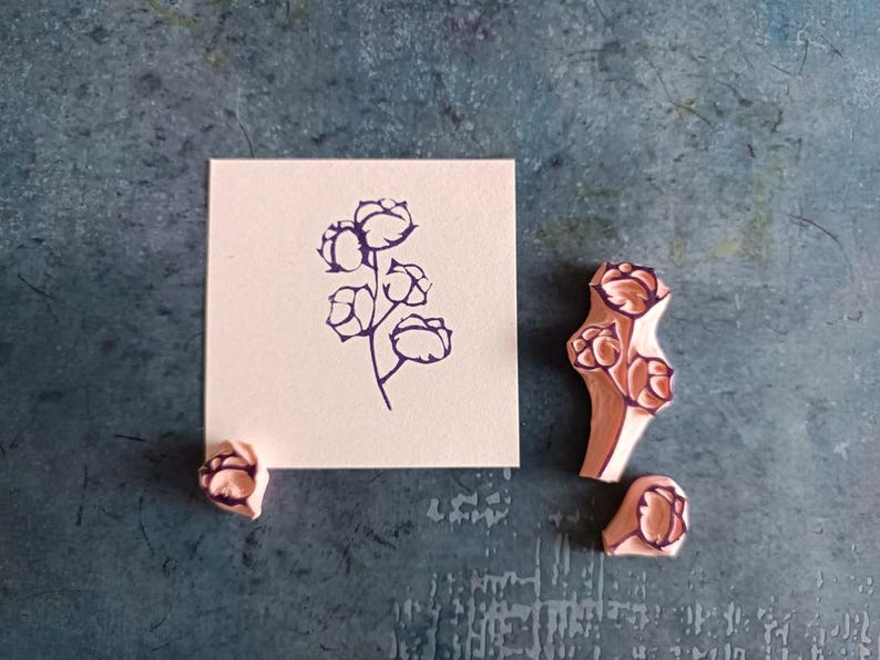 May include: A square, off-white card with a dark purple floral design, alongside three wooden stamps. The stamps and the design on the card feature a cotton plant motif. The background is a textured, teal-colored surface.