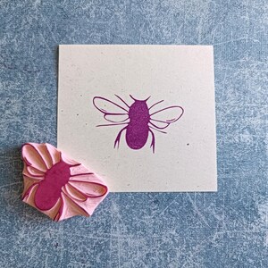 May include: A pink rubber stamp of a bee next to a cream-colored square with a purple bee illustration. The stamp has a detailed bee design, and the printed image shows the same bee. The background is a textured blue surface.