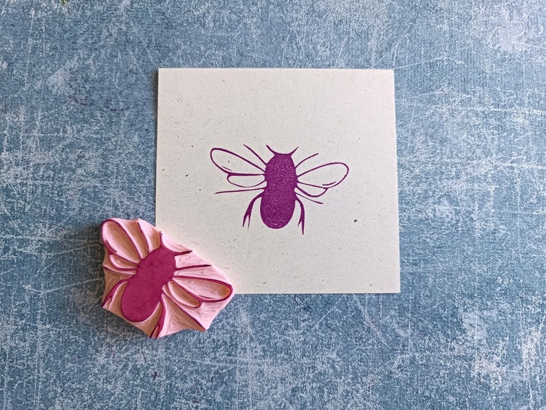 May include: A pink rubber stamp of a bee next to a cream-colored square with a purple bee illustration. The stamp has a detailed bee design, and the printed image shows the same bee. The background is a textured blue surface.