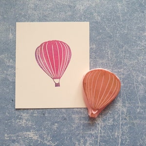 Pode incluir: Um carimbo de borracha rosa de um bal&atilde;o de ar quente. O carimbo est&aacute; em um cart&atilde;o branco com uma impress&atilde;o rosa do bal&atilde;o.