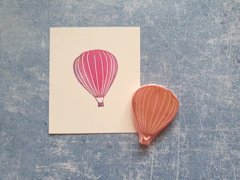 Pode incluir: Um carimbo de borracha rosa de um bal&atilde;o de ar quente. O carimbo est&aacute; em um cart&atilde;o branco com uma impress&atilde;o rosa do bal&atilde;o.
