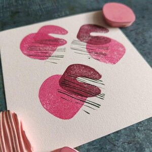 May include: A close-up of a white card with three abstract pink and maroon shapes stamped on it. The shapes are a combination of a heart and a curved line. A pink stamp and a pink stamp pad are also visible.