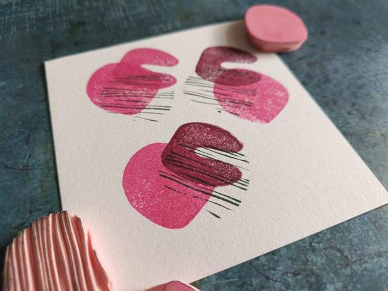 May include: A close-up of a white card with three abstract pink and maroon shapes stamped on it. The shapes are a combination of a heart and a curved line. A pink stamp and a pink stamp pad are also visible.