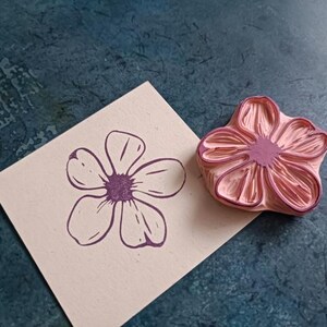 Pode incluir: Um carimbo de borracha rosa com um desenho de flor detalhado. O carimbo &eacute; usado para criar uma impress&atilde;o de flor roxa em um cart&atilde;o bege.