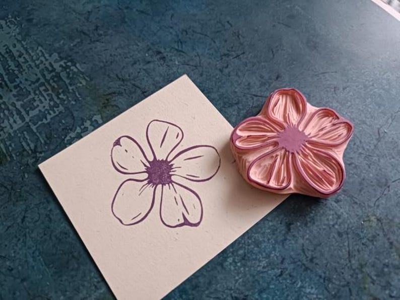Pode incluir: Um carimbo de borracha rosa com um desenho de flor detalhado. O carimbo &eacute; usado para criar uma impress&atilde;o de flor roxa em um cart&atilde;o bege.