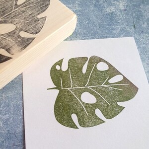 May include: A wooden stamp with a carved monstera leaf design next to a print of the same leaf in green ink on white paper. The stamp has a rectangular shape with a rounded edge. The leaf print has several holes.