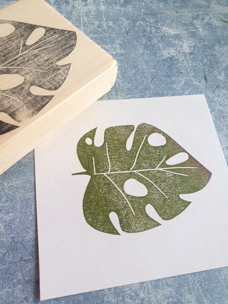 May include: A wooden stamp with a carved monstera leaf design next to a print of the same leaf in green ink on white paper. The stamp has a rectangular shape with a rounded edge. The leaf print has several holes.