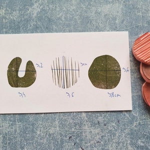May include: A white card displays three green ink stamp impressions: a U-shape, a circle with vertical lines, and an irregular circle. Dimensions are marked in centimeters. Three pink rubber stamps are stacked on the right.