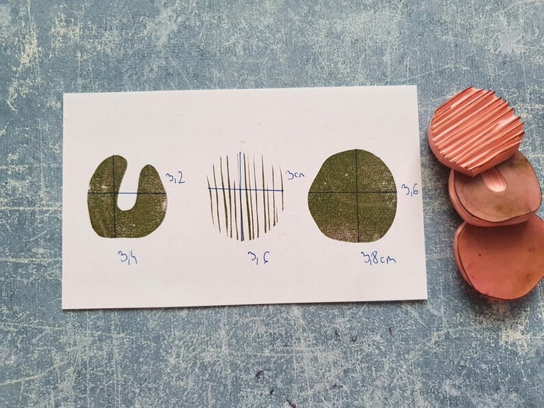 May include: A white card displays three green ink stamp impressions: a U-shape, a circle with vertical lines, and an irregular circle. Dimensions are marked in centimeters. Three pink rubber stamps are stacked on the right.