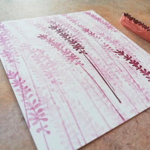 May include: A close-up of a white card with a pink and purple floral design created with a rubber stamp. The stamp is visible in the upper right corner of the image.