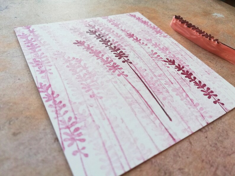 May include: A close-up of a white card with a pink and purple floral design created with a rubber stamp. The stamp is visible in the upper right corner of the image.