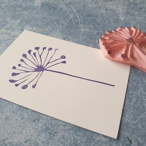 May include: A purple rubber stamp of a stylized dandelion with a long stem. The stamp is on a white card, and a pink rubber stamp of a dandelion is lying next to it.
