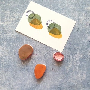 May include: A set of four polymer clay earrings in shades of green, orange, and pink. The earrings are shaped like abstract geometric shapes with a circle design.