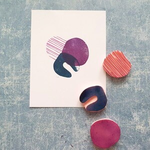 May include: A white card with abstract shapes in purple, teal, and pink. Three rubber stamps with similar shapes are next to the card. The stamps are pink, teal, and purple. The background is a textured blue surface.