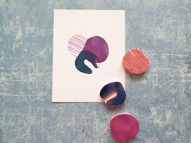 May include: A white card with abstract shapes in purple, teal, and pink. Three rubber stamps with similar shapes are next to the card. The stamps are pink, teal, and purple. The background is a textured blue surface.
