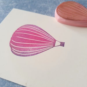 Pode incluir: Um carimbo de borracha rosa de um bal&atilde;o de ar quente com uma cesta. O carimbo est&aacute; em um cart&atilde;o branco.