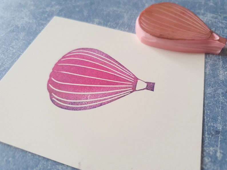 Pode incluir: Um carimbo de borracha rosa de um bal&atilde;o de ar quente com uma cesta. O carimbo est&aacute; em um cart&atilde;o branco.